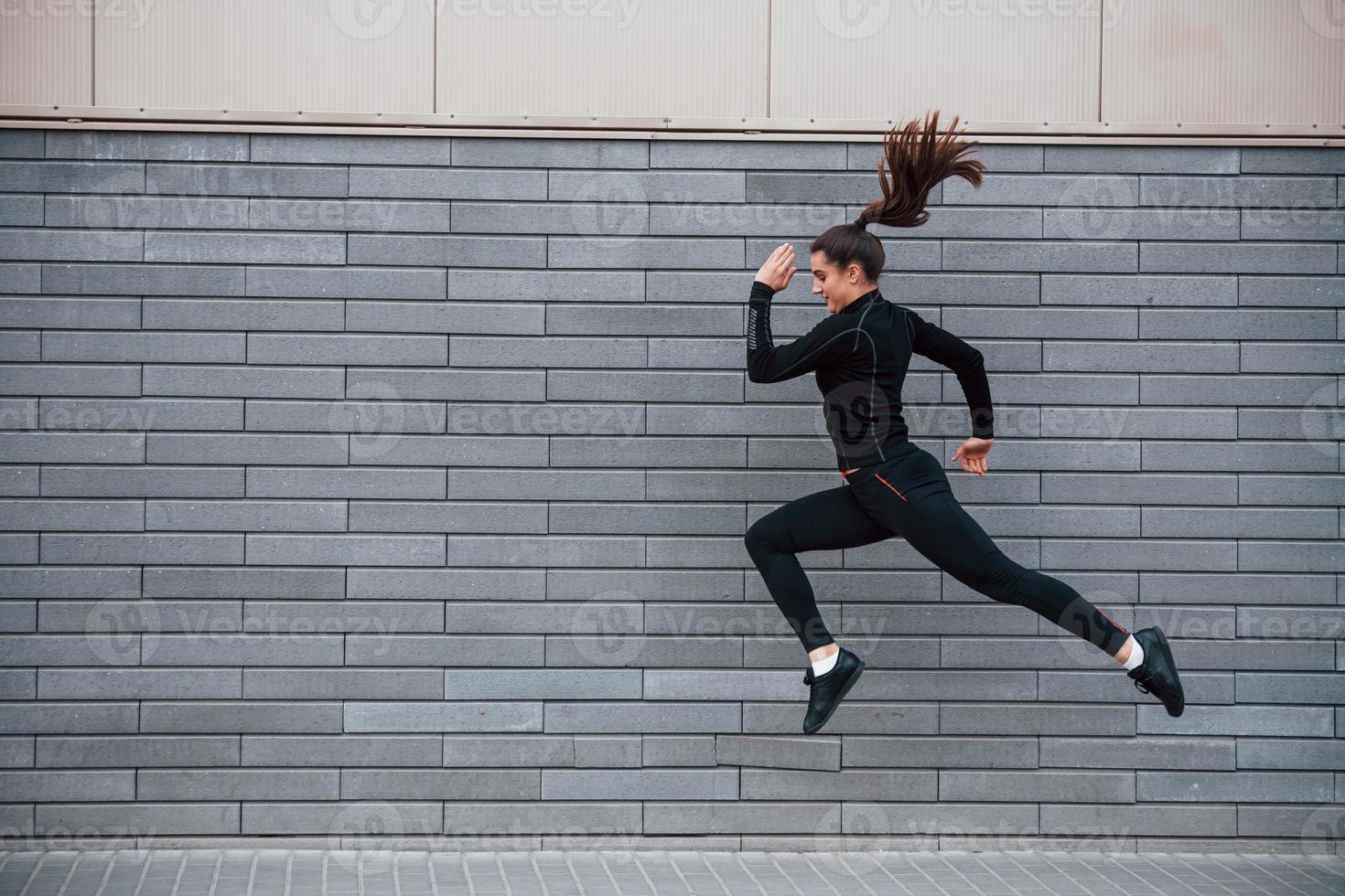 Young sportive girl in black sportswear running outdoors near gray wall photo