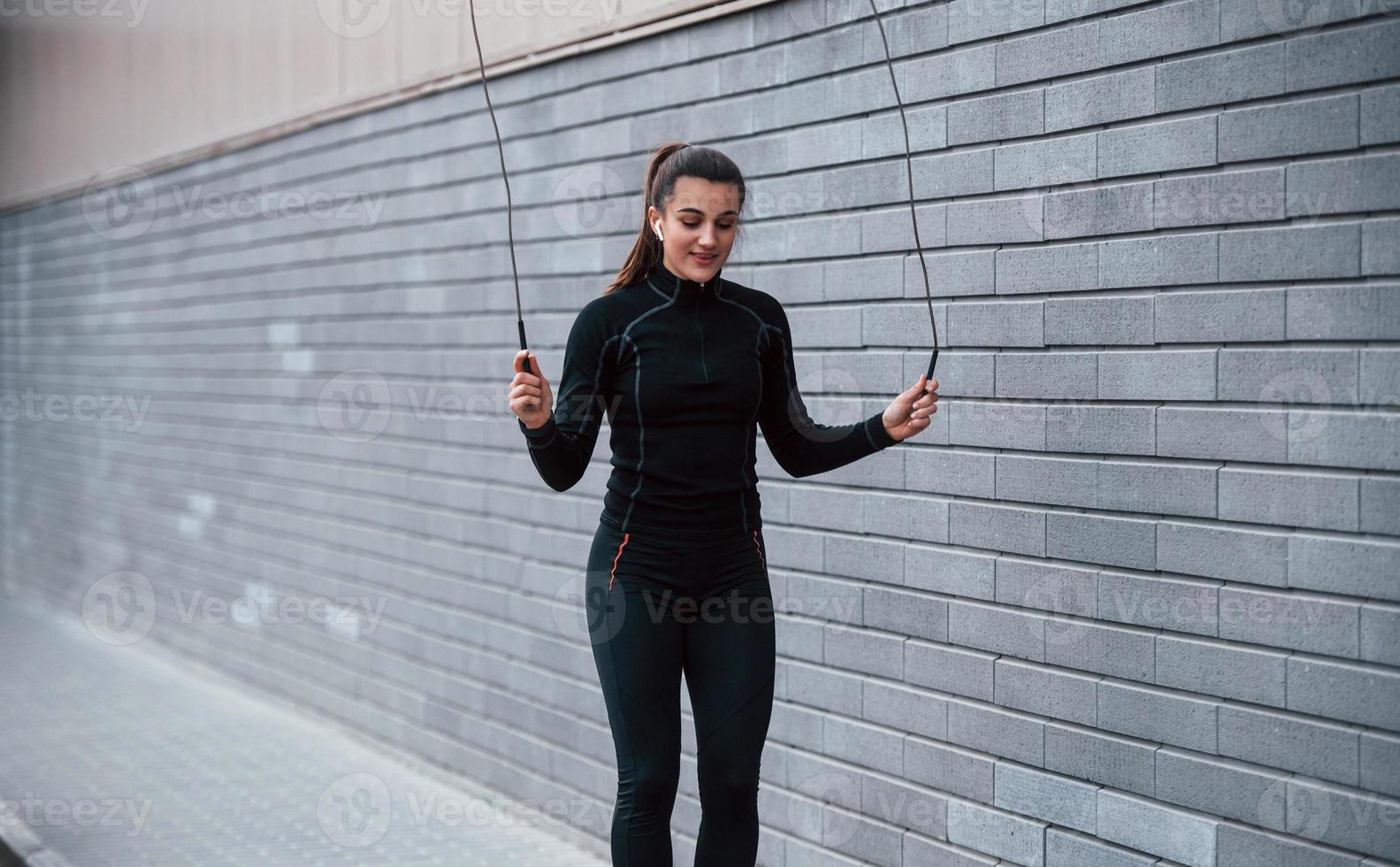 Young sportive girl in black sportswear practicing with jump rope outdoors near gray wall photo