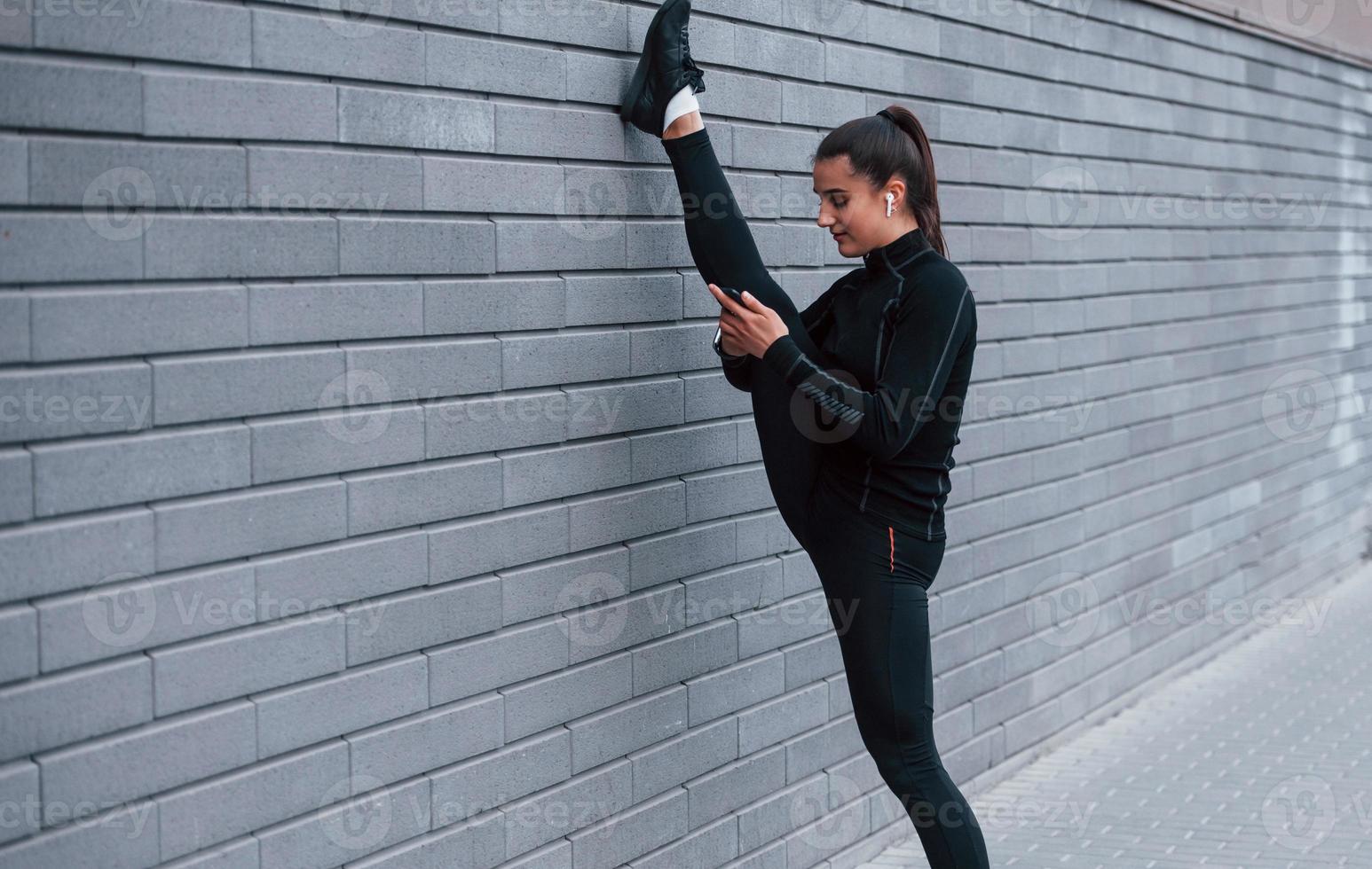 Young sportive girl with phone in black sportswear doing legs stretching outdoors by using gray wall photo