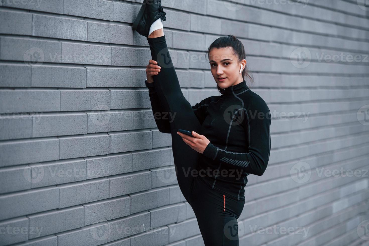 Young sportive girl with phone in black sportswear doing legs stretching outdoors by using gray wall photo