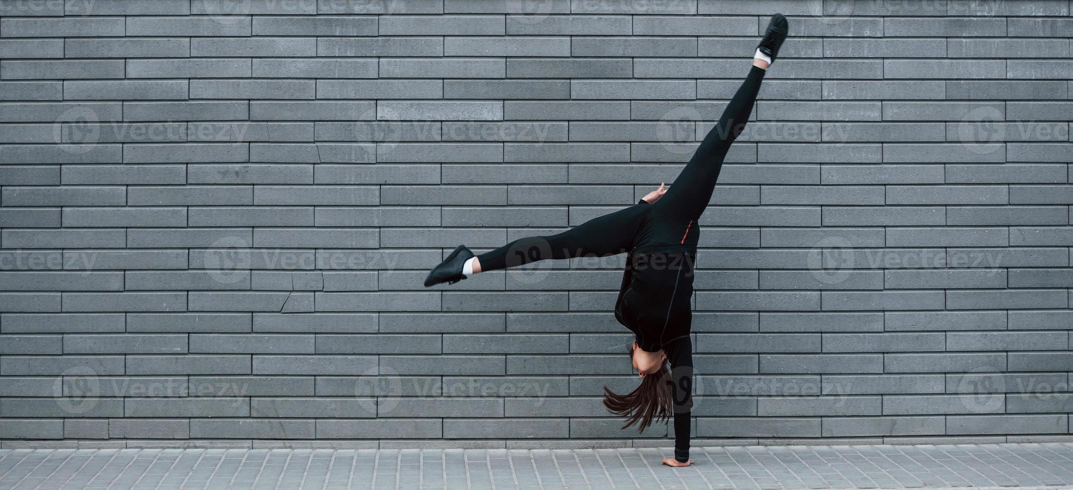 Young sportive girl in black sportswear doing hard handstand exercises outdoors near gray wall photo