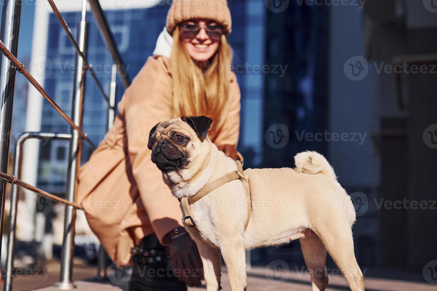Woman in warm clothes sitting and have fun with her little pug dog near business building that on background photo
