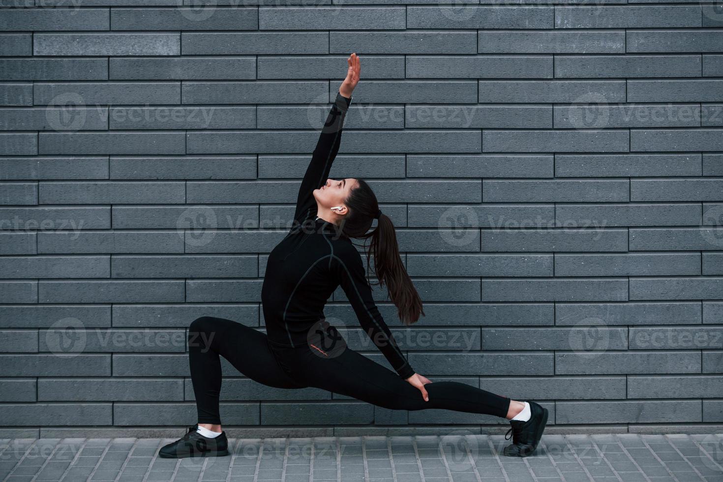 joven deportista con ropa deportiva negra al aire libre haciendo ejercicios de yoga cerca de la pared gris foto