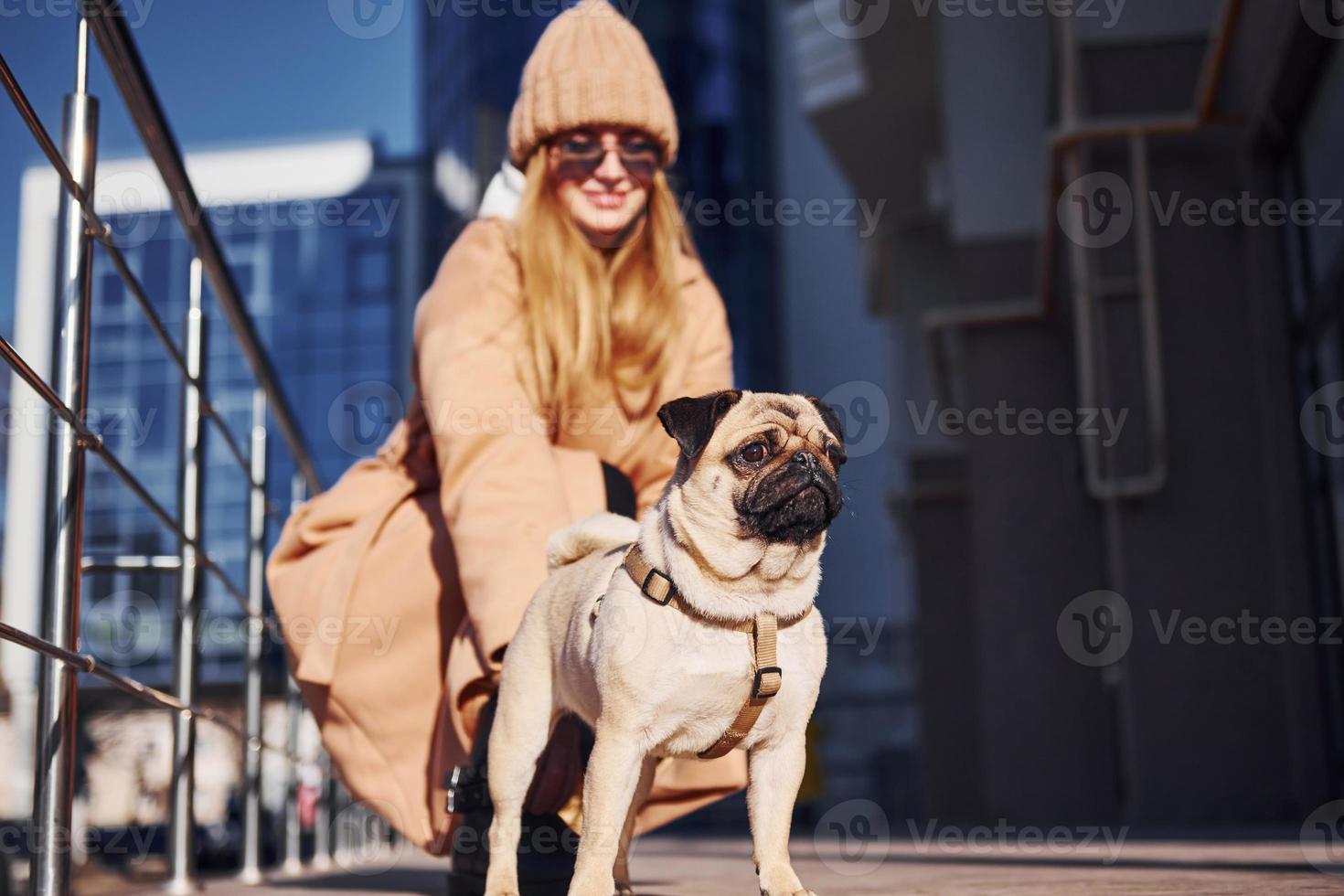 Woman in warm clothes sitting and have fun with her little pug dog near business building that on background photo