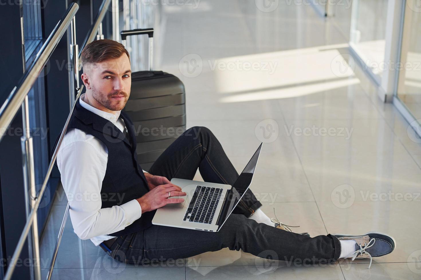Young male passenger in elegant formal clothes sitting with laptop in the airport hall photo