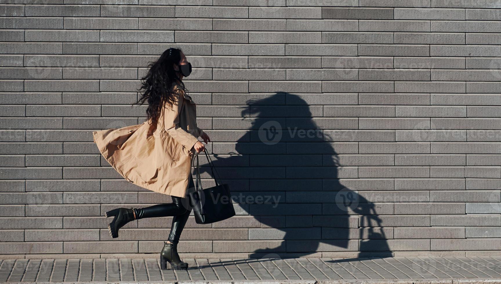 en máscara protectora. hermosa morena con cabello rizado y ropa negra corriendo al aire libre cerca de la pared foto