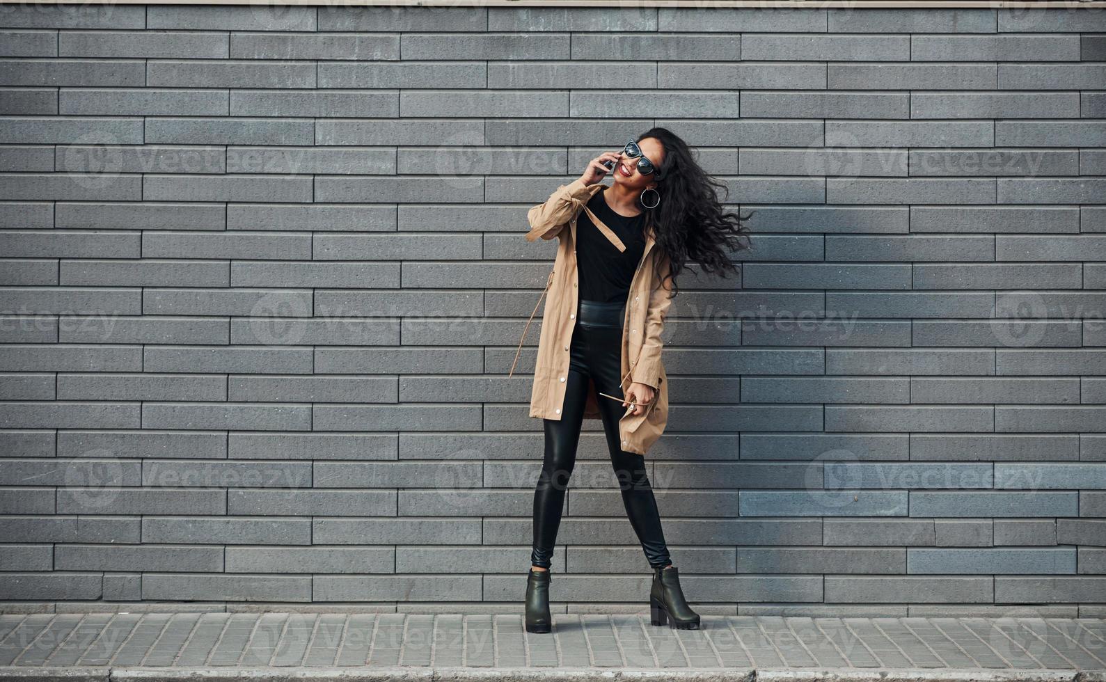 Beautiful brunette with curly hair and in black clothes talking by phone outdoors near wall photo