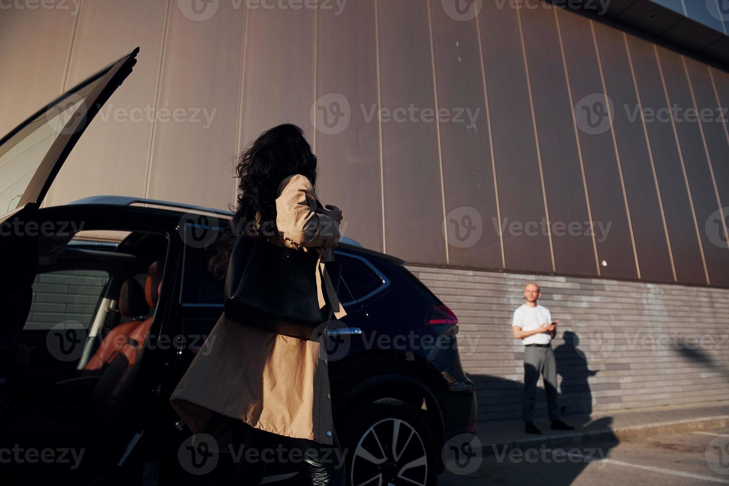 Man at background. Young woman in black leggins walks out from her modern car photo