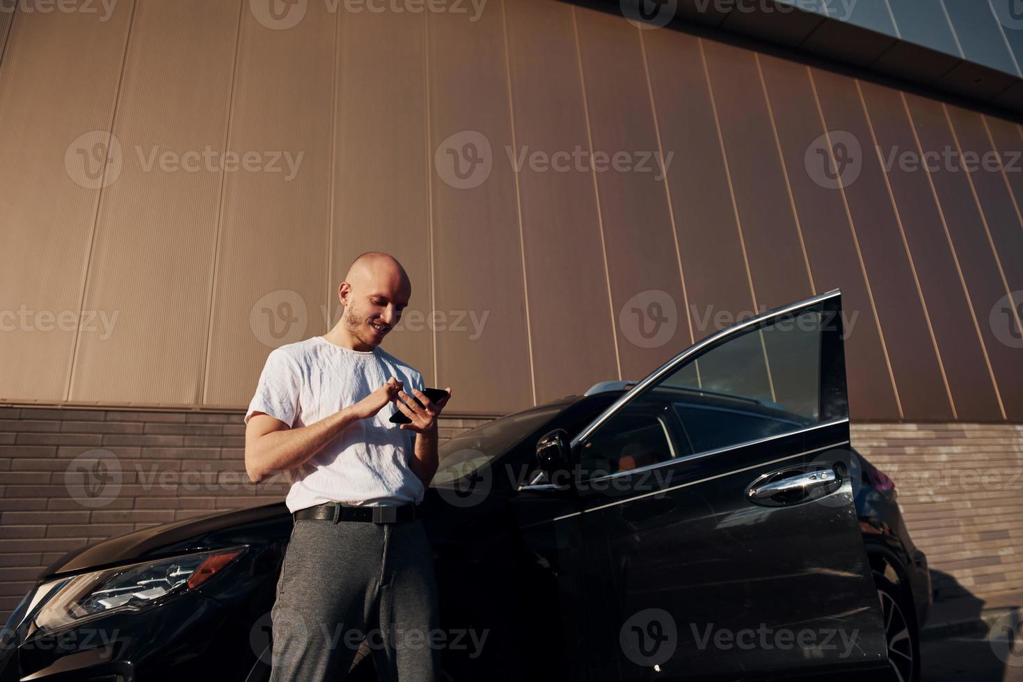 Bald man standing near his modern black car outdoors at daytime with phone in hands photo