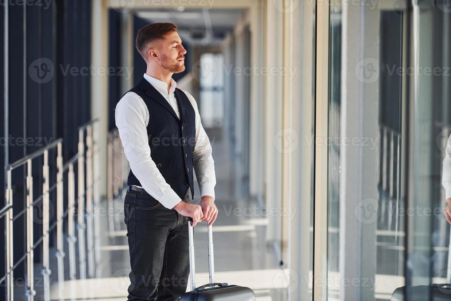 Young male passenger in elegant formal clothes is in the airport hall standing with baggage photo