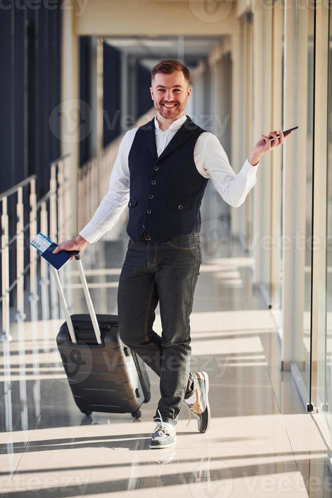 el pasajero masculino con ropa formal elegante está en la sala del aeropuerto con equipaje, boletos y teléfono foto