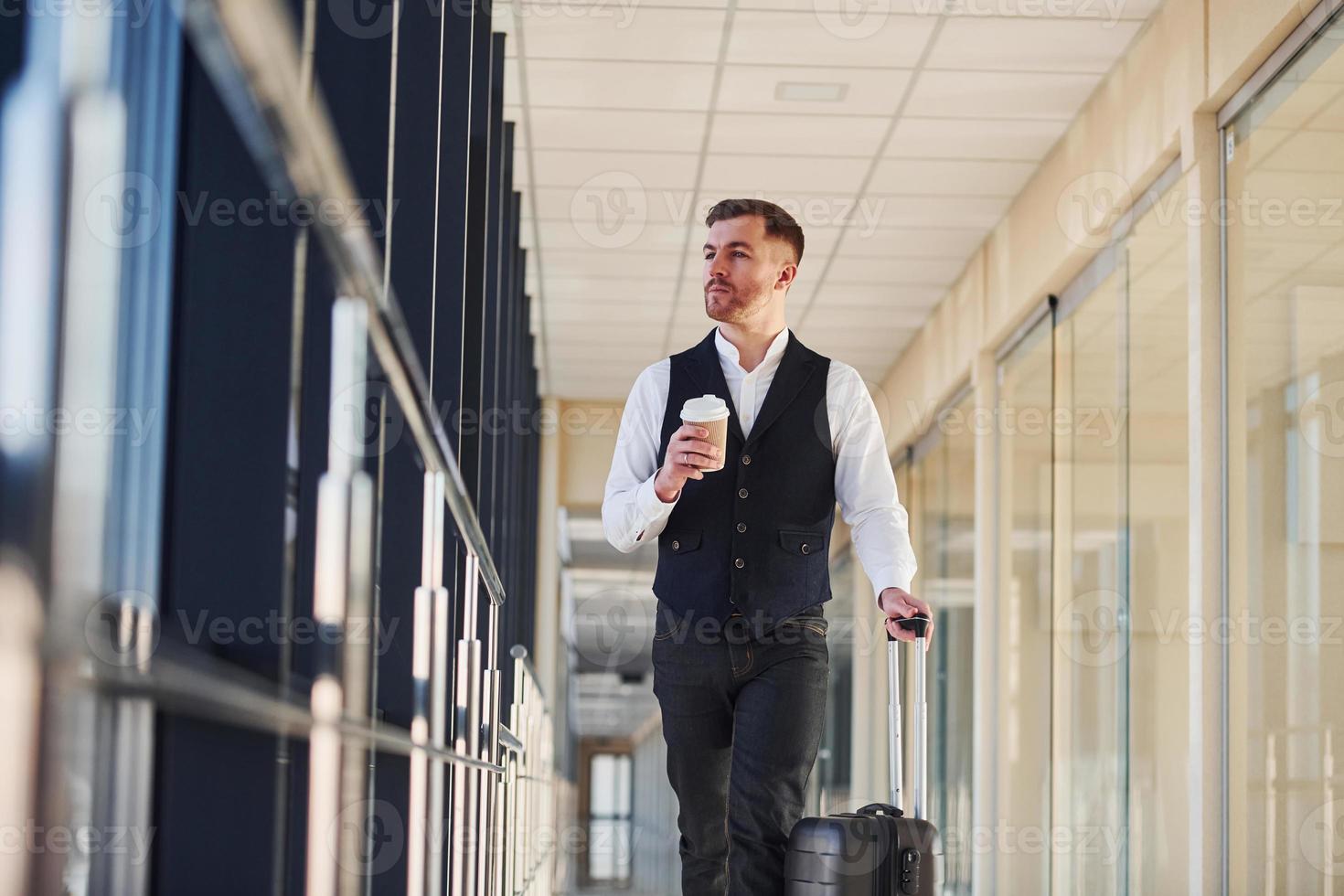 un joven pasajero vestido con ropa elegante y formal está en la sala del aeropuerto caminando con equipaje y una taza de bebida foto