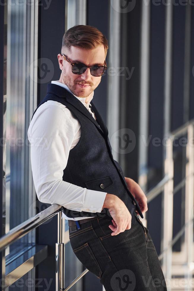 Man in elegant clothes and sunglasses is standing indoors and posing for the camera photo