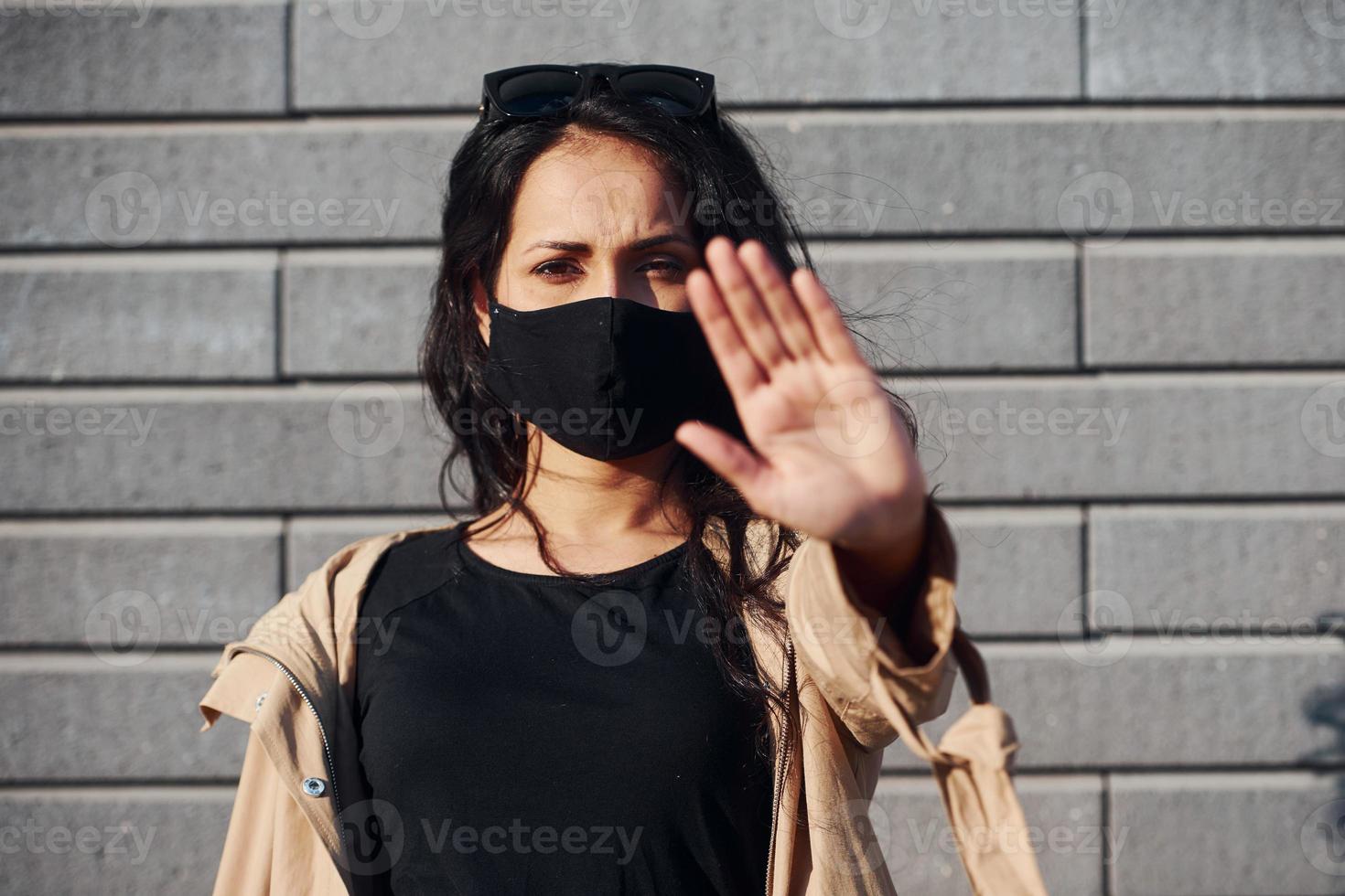 mostrando gesto de parada con la mano. en máscara protectora. hermosa morena con cabello rizado y ropa negra al aire libre cerca de la pared foto