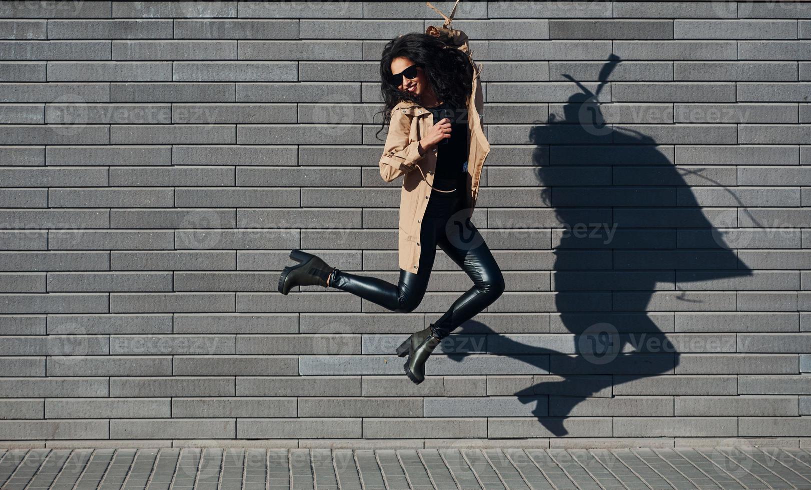 Beautiful brunette with curly hair and in black clothes jumping and having fun outdoors near wall photo