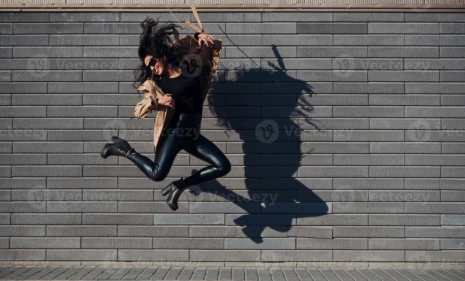 hermosa morena con el pelo rizado y vestida de negro saltando y divirtiéndose al aire libre cerca de la pared foto