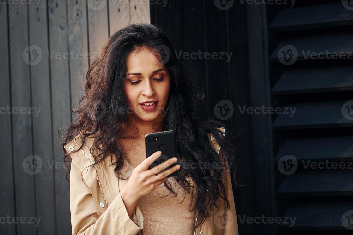 Woman with black curly hair standing against black wooden building exterior and using phone photo