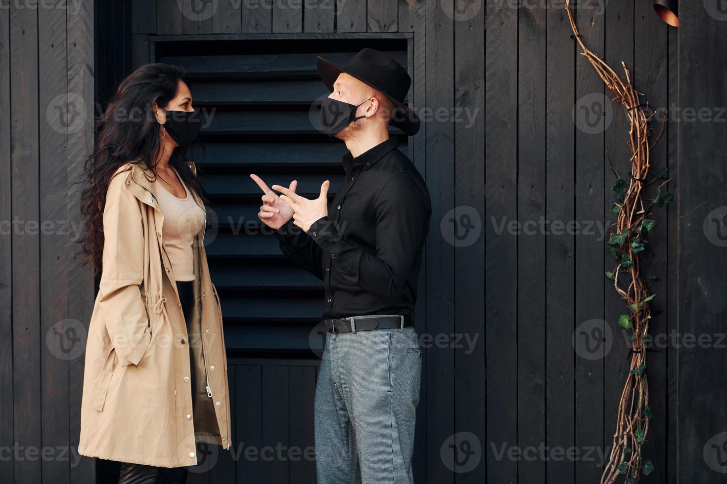 Woman with black curly hair standing against black wooden building exterior with her man in hat photo