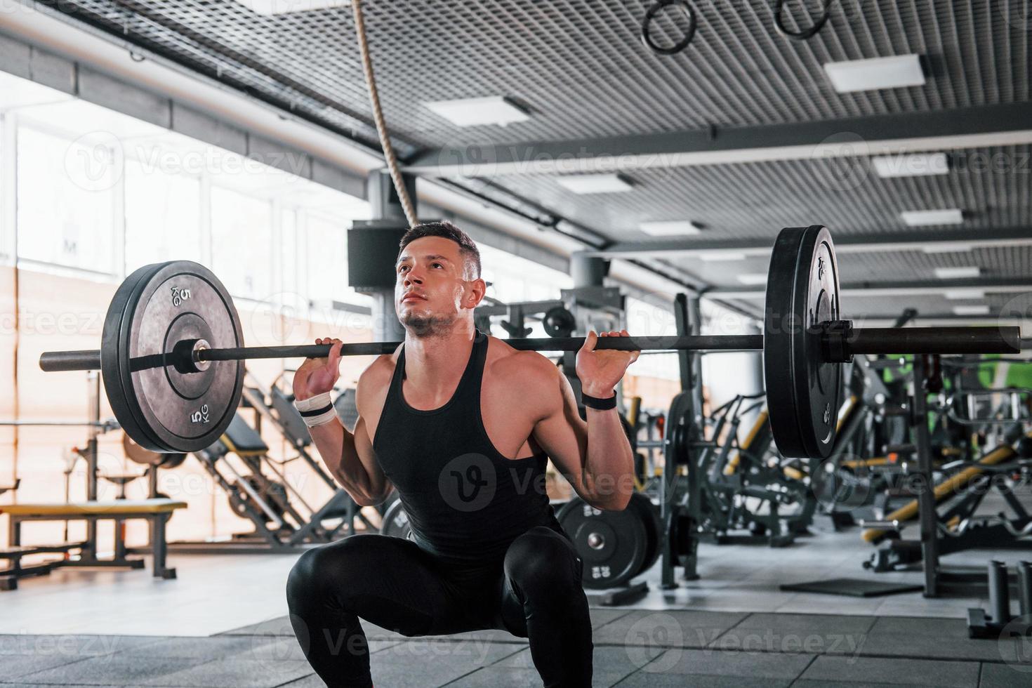 Lifting barbell. Young sportive strong man in black wear have workout day in gym photo