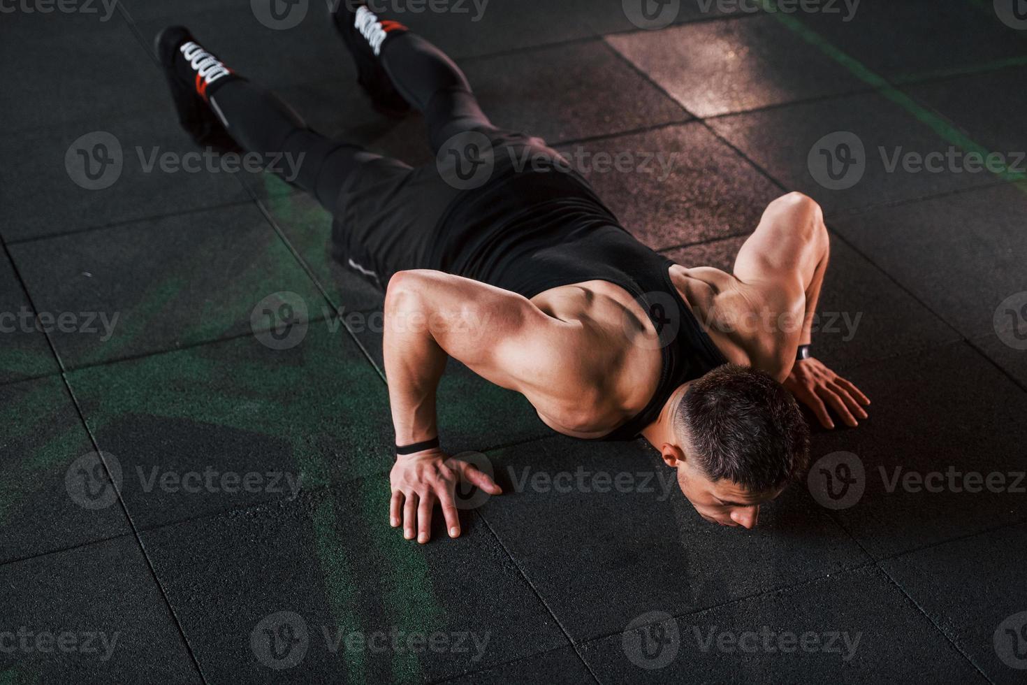 Doing push-ups. Young sportive strong man in black wear have workout day in gym photo