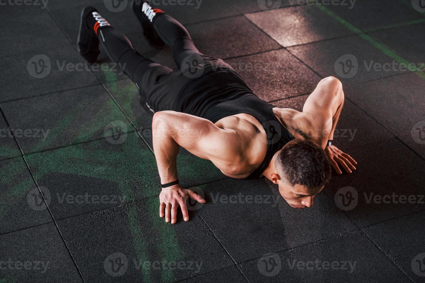 Doing push-ups. Young sportive strong man in black wear have workout day in gym photo