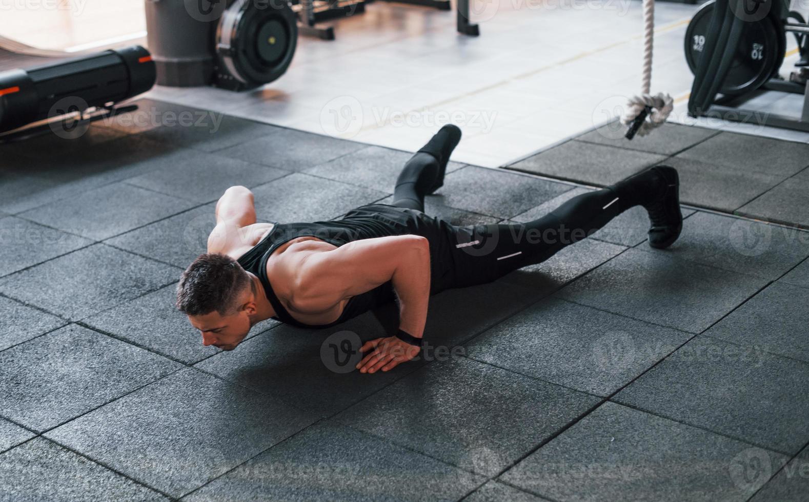 Doing push-ups. Young sportive strong man in black wear have workout day in gym photo