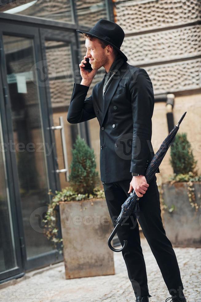 Talking by phone. Elegant young man in black hat and clothes with umbrella in hands outdoors in the city photo