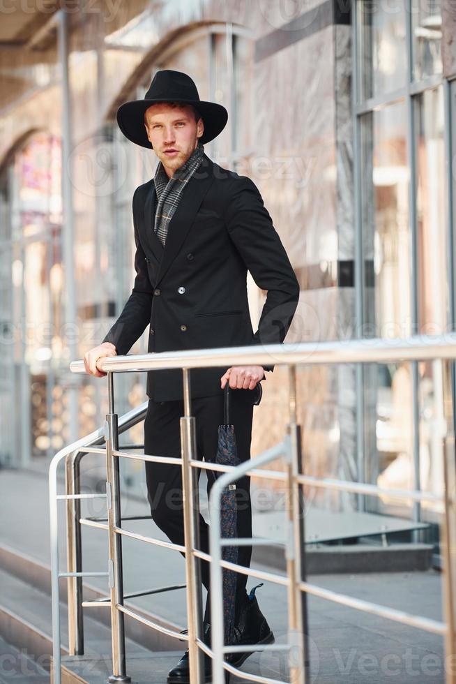 joven elegante con sombrero negro y paraguas al aire libre en la ciudad foto