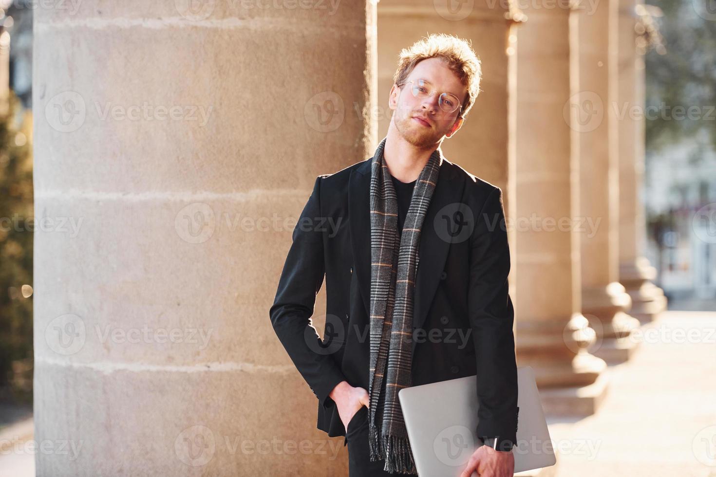 With laptop in hands. Elegant young man in glasses adn in formal classy clothes outdoors in the city photo