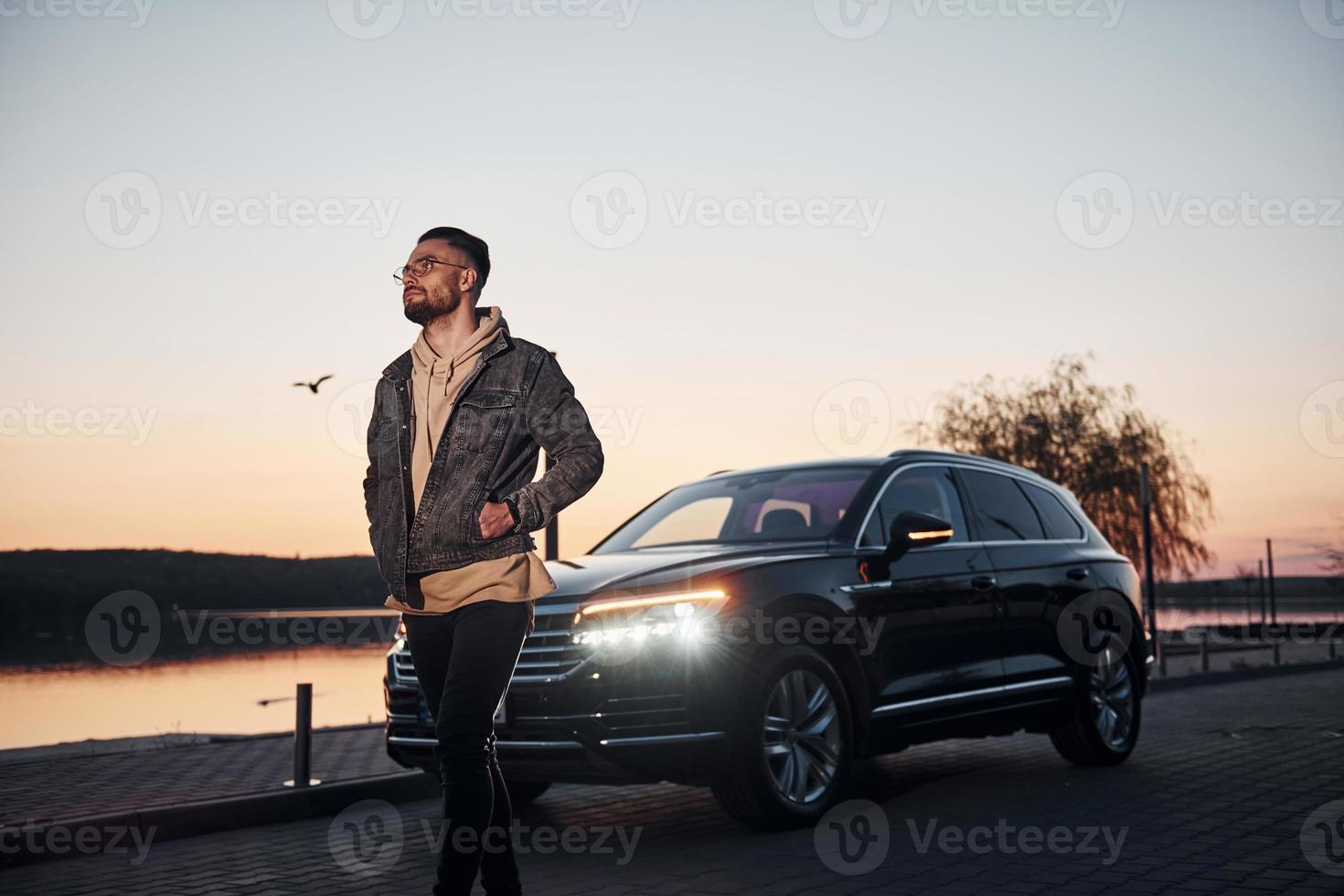 Handsome unshaved man in fashionable clothes walks near his black car at evening time photo