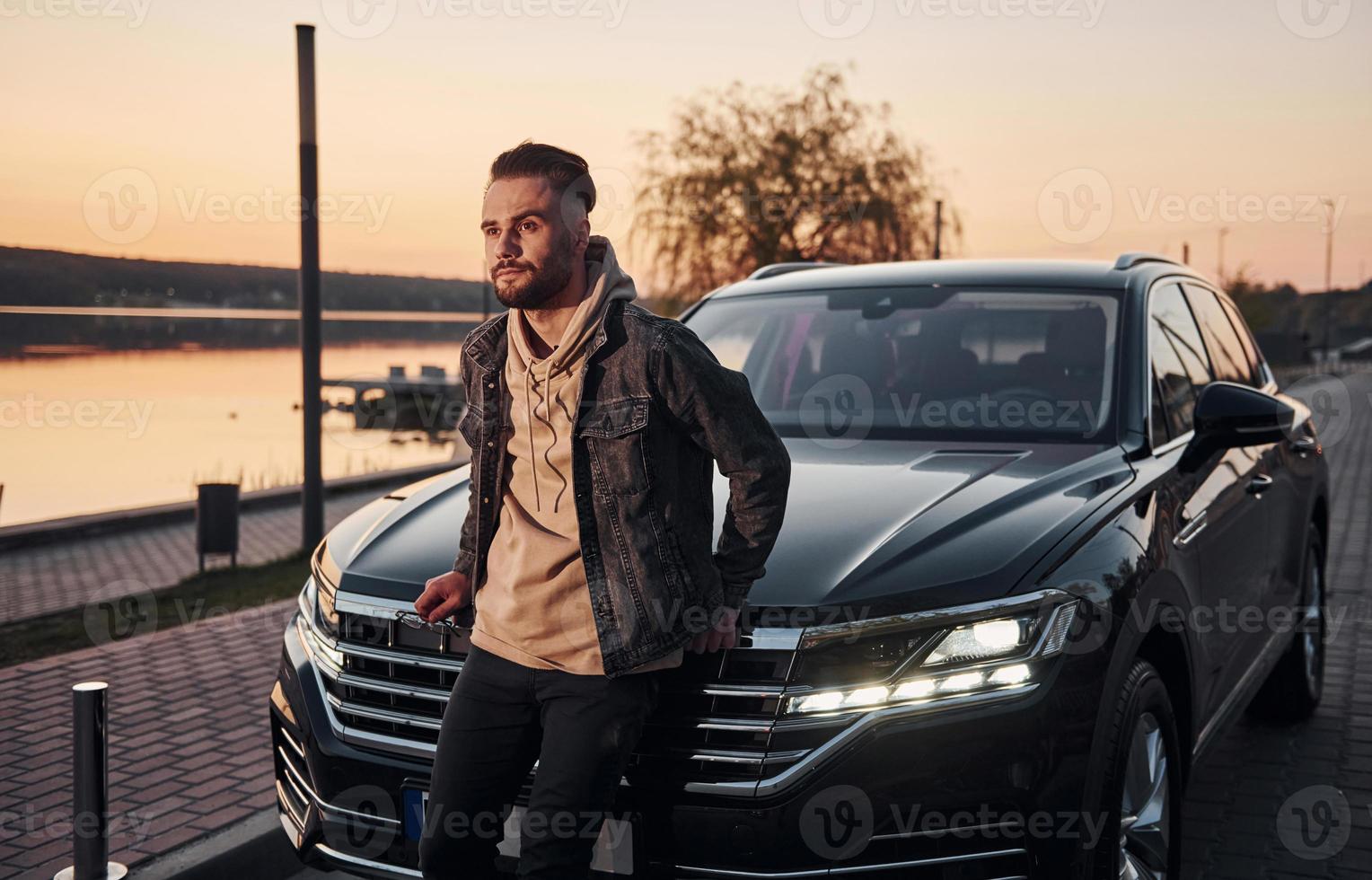 Handsome unshaved man in fashionable clothesleaning on his his black car photo