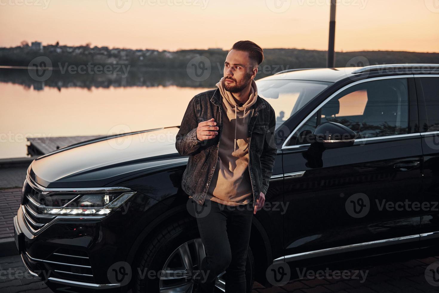 Handsome unshaved man in fashionable clothes standing near his black car and smoking photo