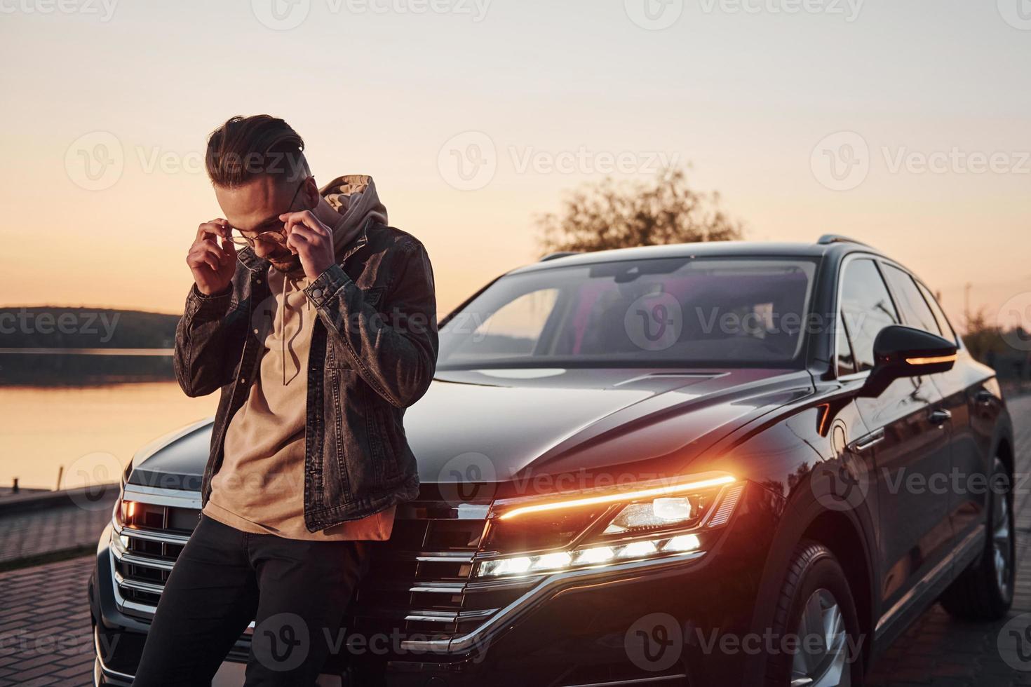 Handsome unshaved man in fashionable clothesleaning on his his black car photo