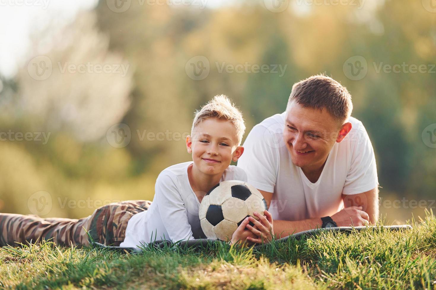With soccer ball. Father with his son lying down on the ground together outdoors near forest at daytime photo