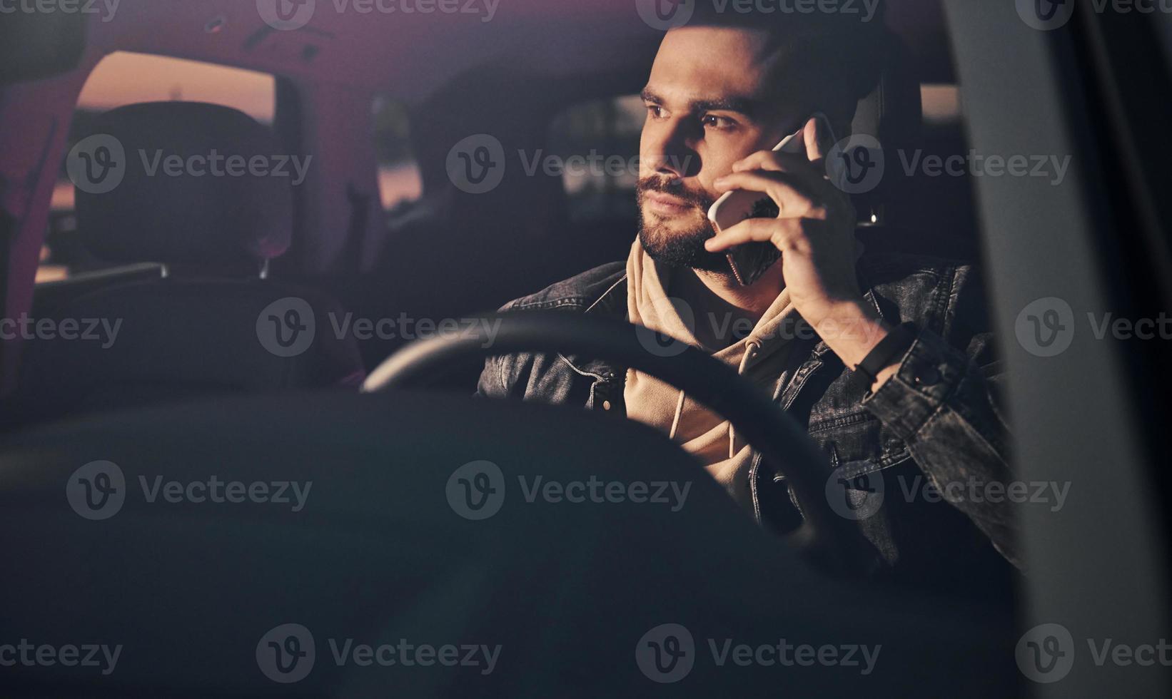 Front view of handsome unshaved man in fashionable clothes that riding his automobile and talks by phone photo