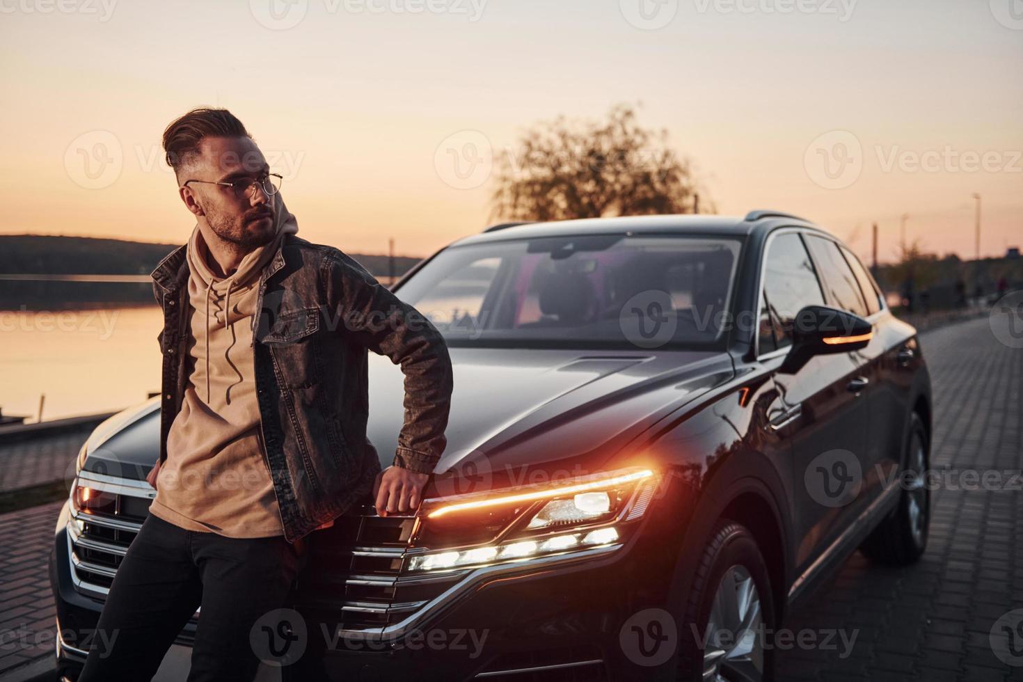 Handsome unshaved man in fashionable clothesleaning on his his black car photo