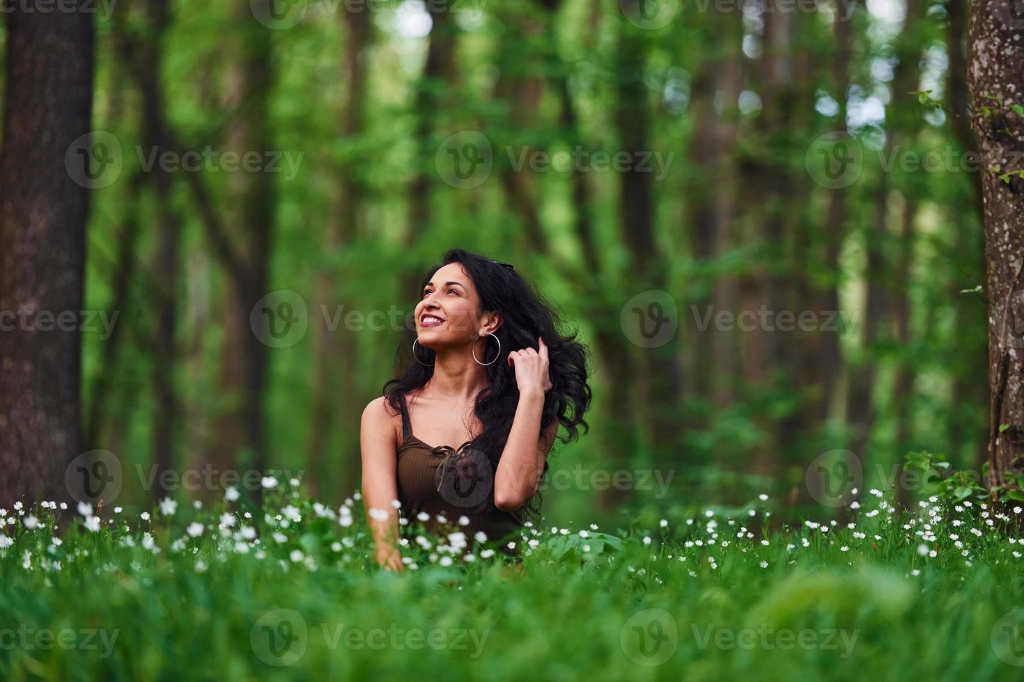 Positive brunette in casual clothes sitting in the forest at daytime photo