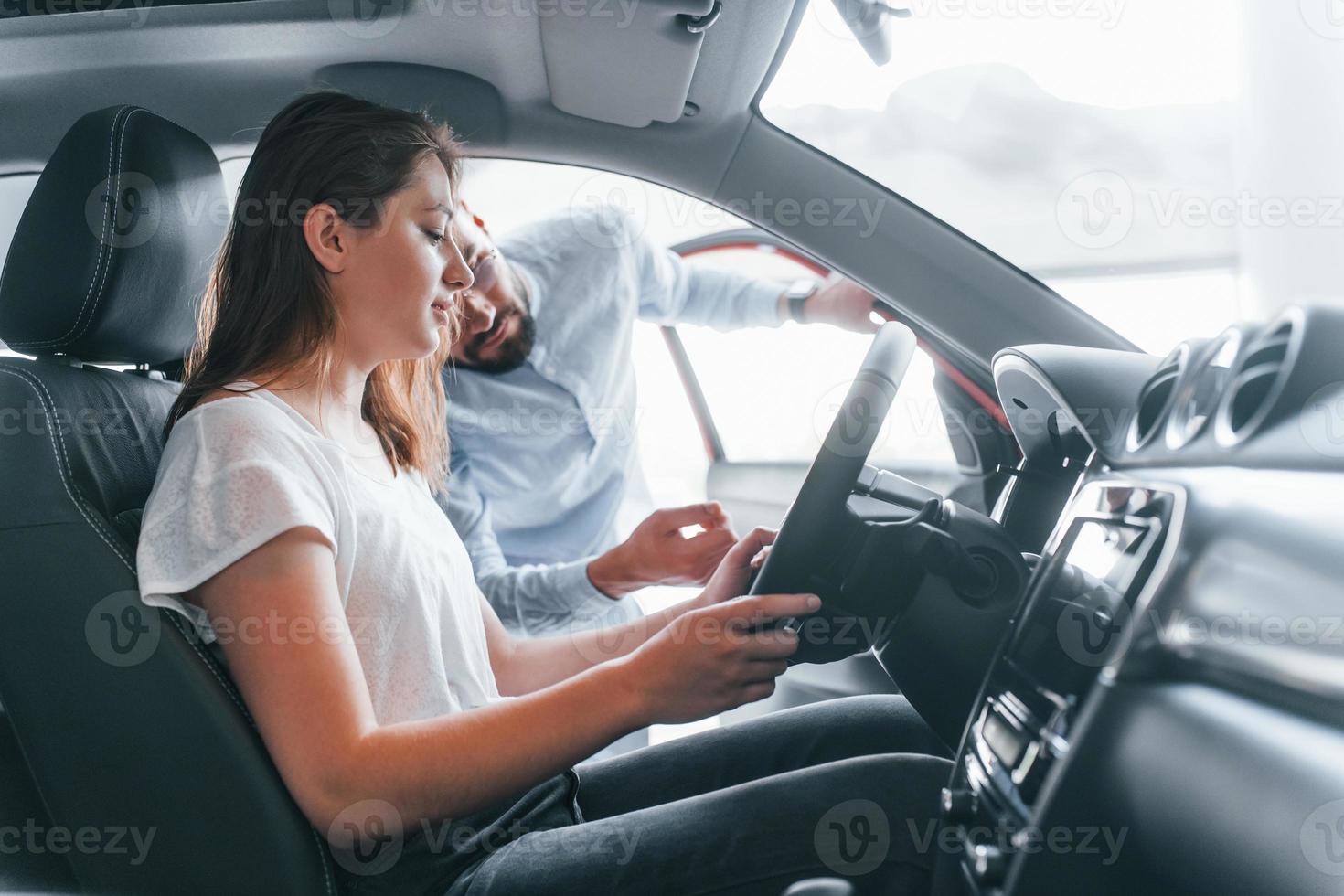 vista lateral de una joven positiva que tiene una visita en autosalon y elige auto. gerente ayudándola foto