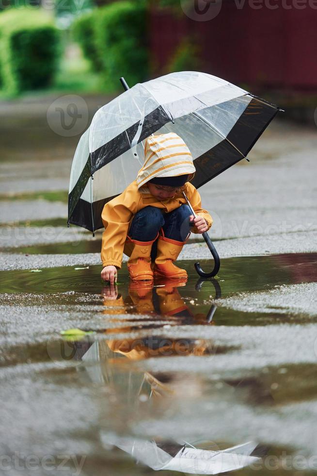 niño con capa impermeable amarilla, botas y paraguas jugando al aire libre después de la lluvia foto