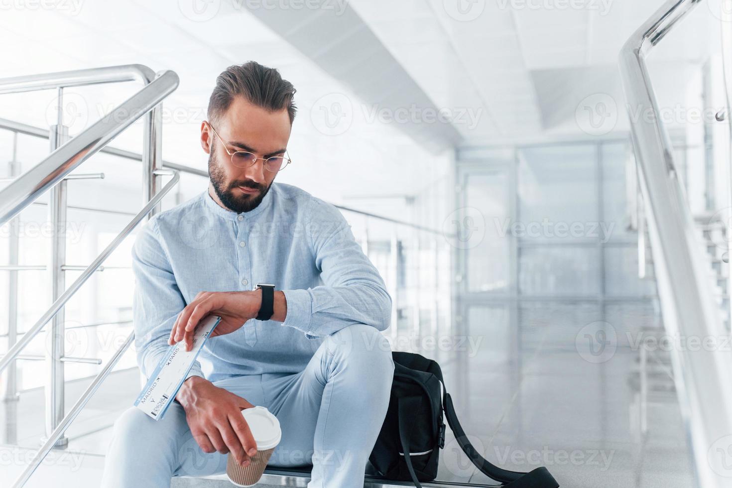 Holding plane tickets in hand. Sitting on stairs and waiting. Young passenger in formal clothes indoors in the office at daytime photo