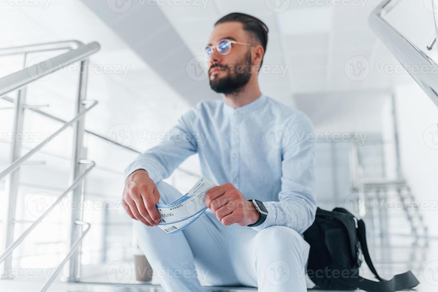 Holding plane tickets in hand. Sitting on stairs and waiting. Young passenger in formal clothes indoors in the office at daytime photo