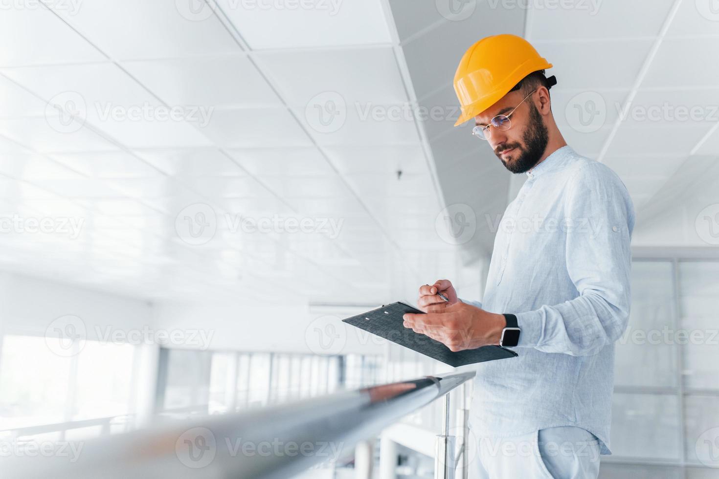 Holding notepad. Engineer in white clothes and orange protective hard hat standing and working indoors photo