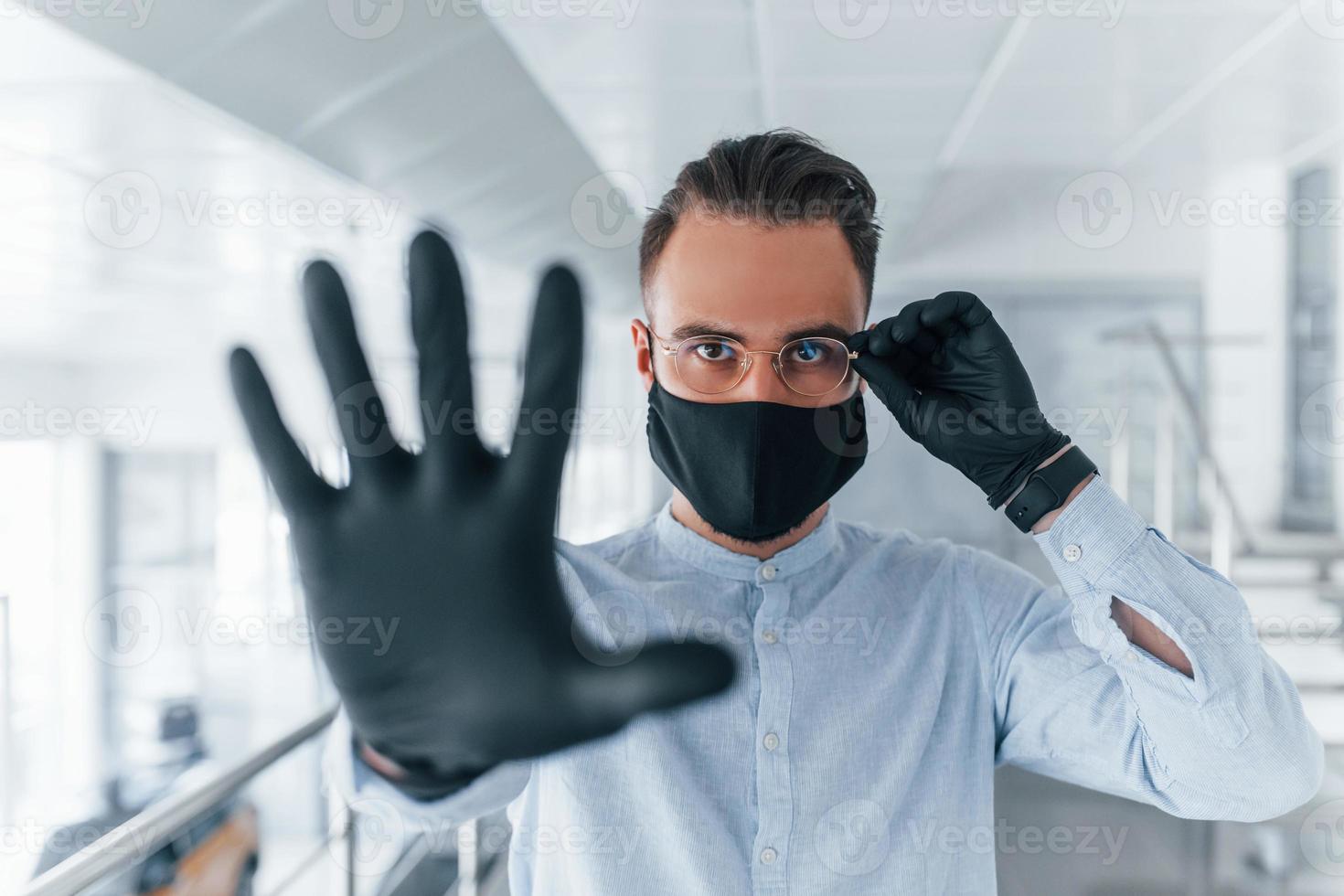 Showing stop gesture by hand. In protective mask and gloves. Young handsome man in formal clothes indoors in the office at daytime photo