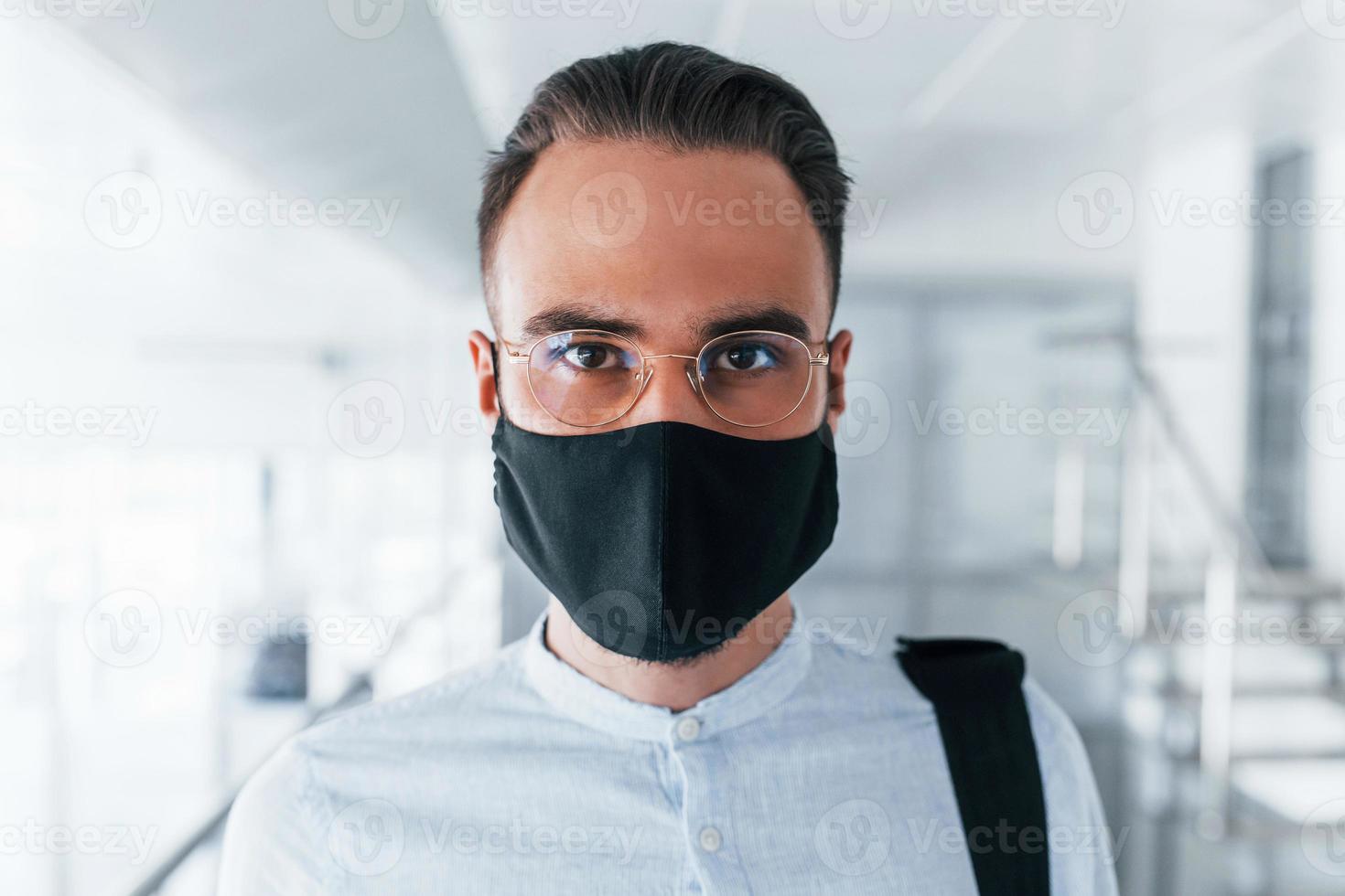 en máscara protectora y guantes. joven apuesto con ropa formal en el interior de la oficina durante el día foto