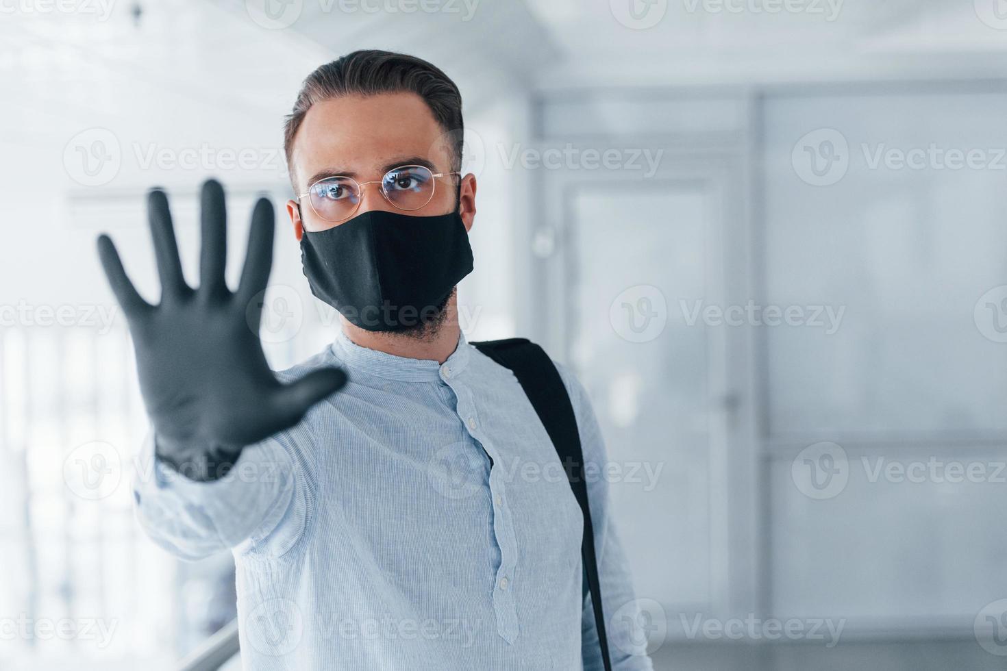 Showing stop gesture by hand. In protective mask and gloves. Young handsome man in formal clothes indoors in the office at daytime photo