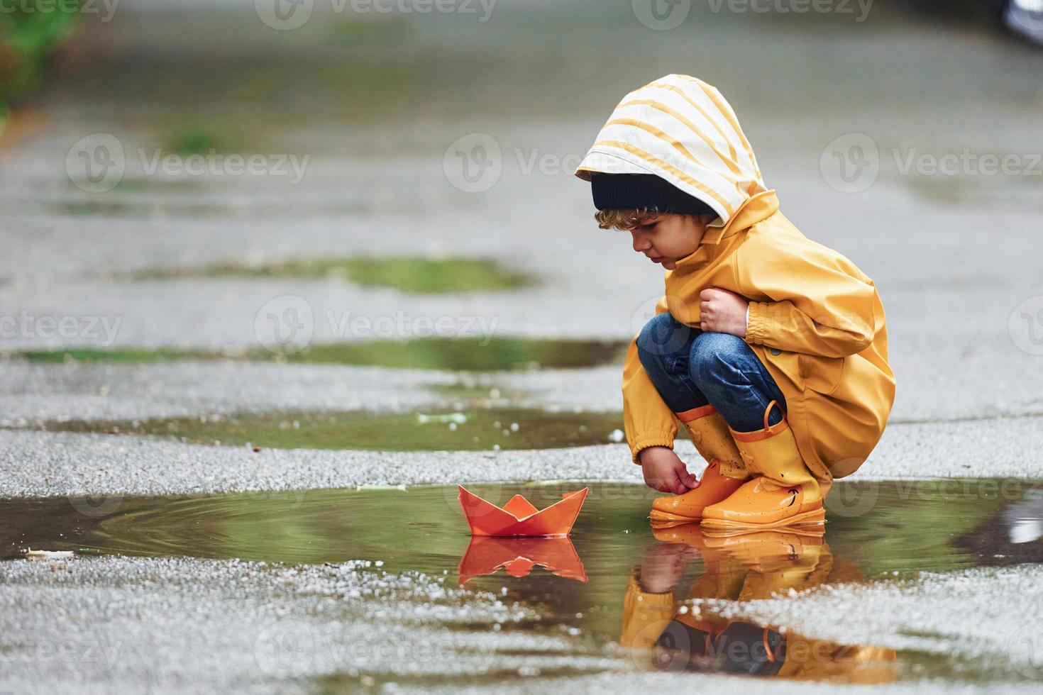 Kid in yellow waterproof cloak and boots playing with paper handmade boat toy outdoors after the rain photo