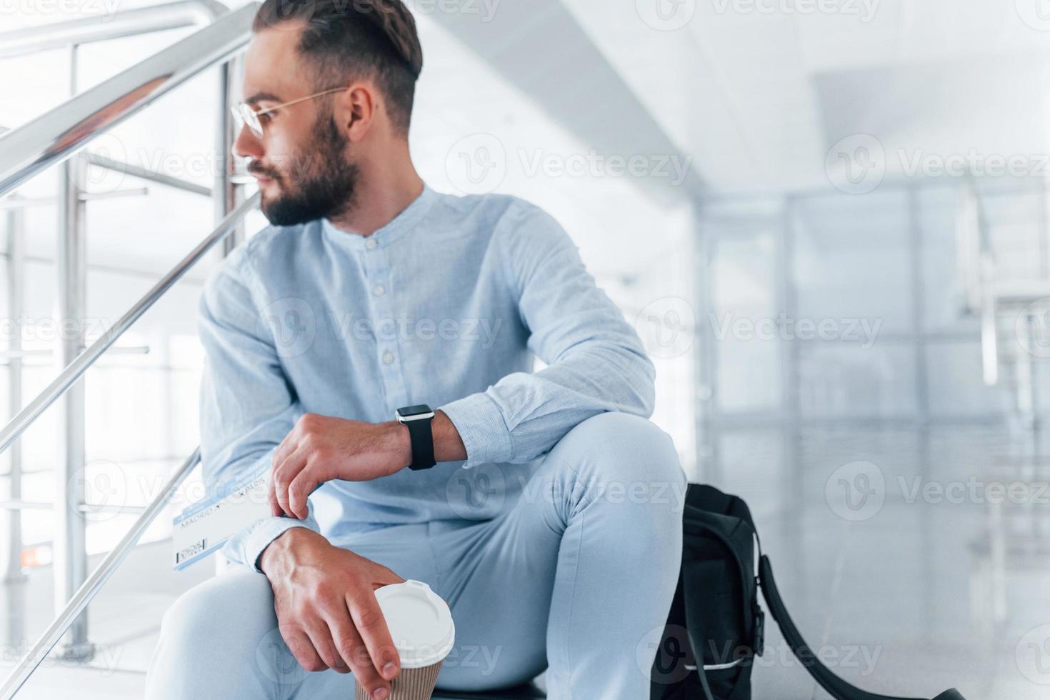 Holding plane tickets in hand. Sitting on stairs and waiting. Young passenger in formal clothes indoors in the office at daytime photo