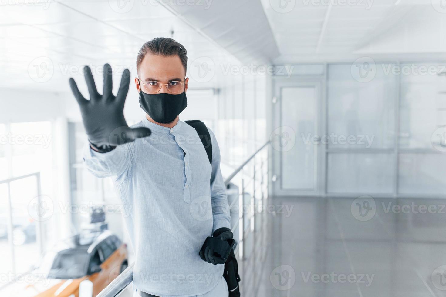 Showing stop gesture by hand. In protective mask and gloves. Young handsome man in formal clothes indoors in the office at daytime photo