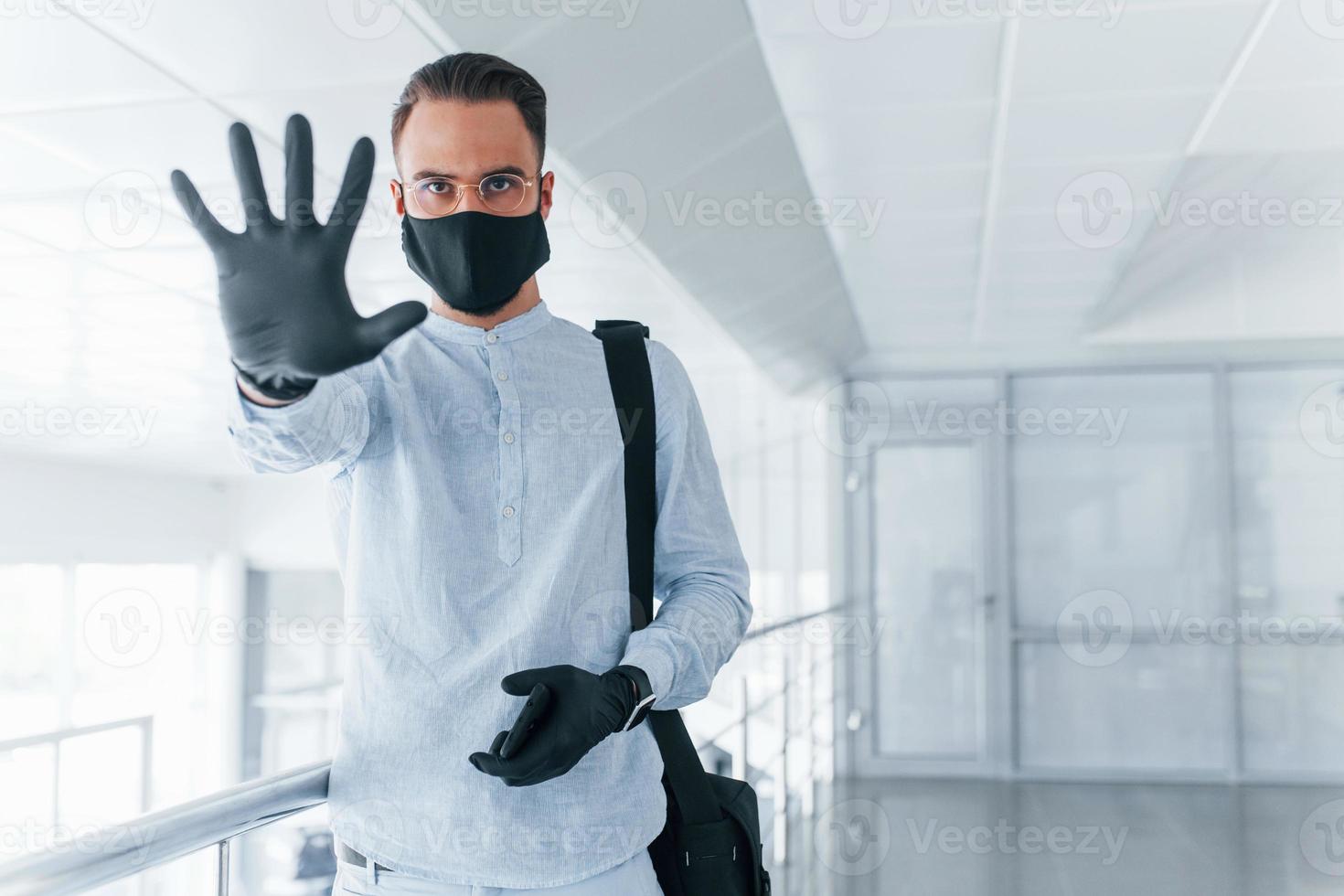 Showing stop gesture by hand. In protective mask and gloves. Young handsome man in formal clothes indoors in the office at daytime photo