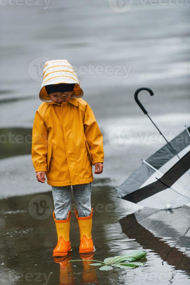 Kid in yellow waterproof cloak, boots and with umbrella playing outdoors after the rain photo